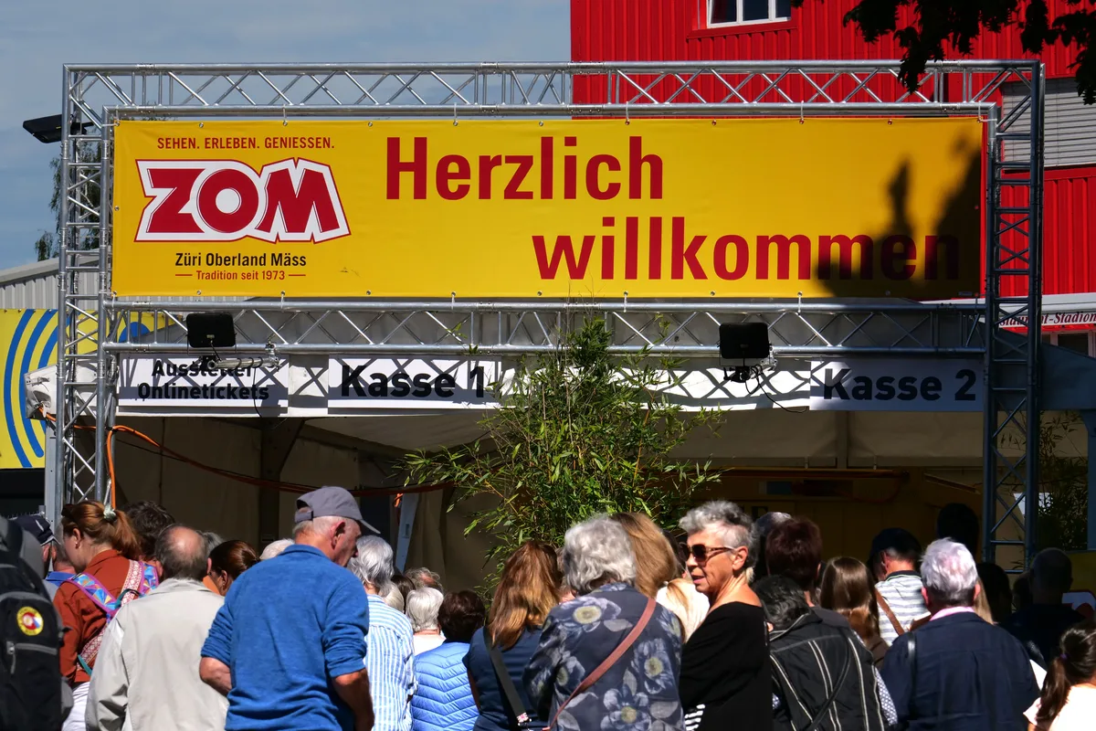Menschen stehen in einer Warteschlange. An deren Ende ist ein Gerüst mit einem gelben Transparent zu sehen. Darauf steht «ZOM: Herzlich willkommen».